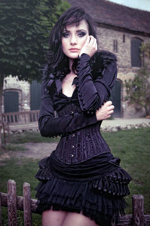 a woman in a black gothicstyle outfit stands in front of a stone building and a wooden fence. she is wearing a black corset, a black skirt, and a black jacket with feather details. she has long dark hair and pale skin, and she is wearing dark makeup. the background is a green lawn and some trees. the lighting is soft and diffused, creating a romantic and slightly eerie atmosphere.