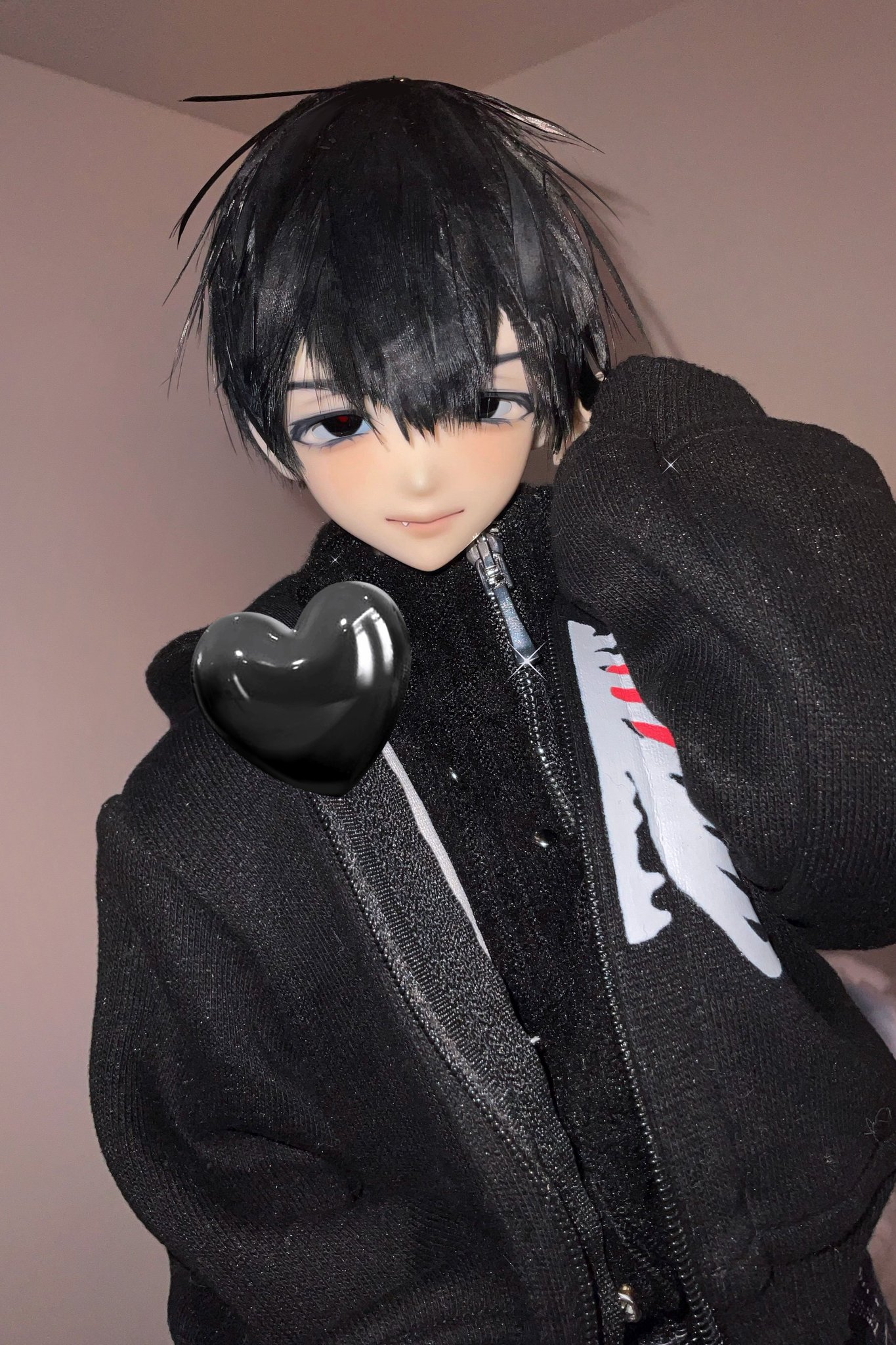 a close up of a doll wearing a black hoodie with a white design on the front, the doll has long black hair and has a black heart sticker on their jacket.