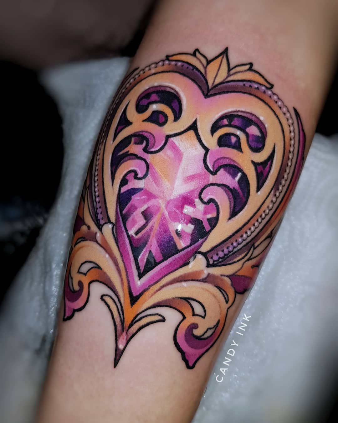 a tattoo of an ornate heartshaped filigree frame encasing a sparkling purple and pink crystal. the tattoo is done in a neotraditional style, with bold colors and intricate details.