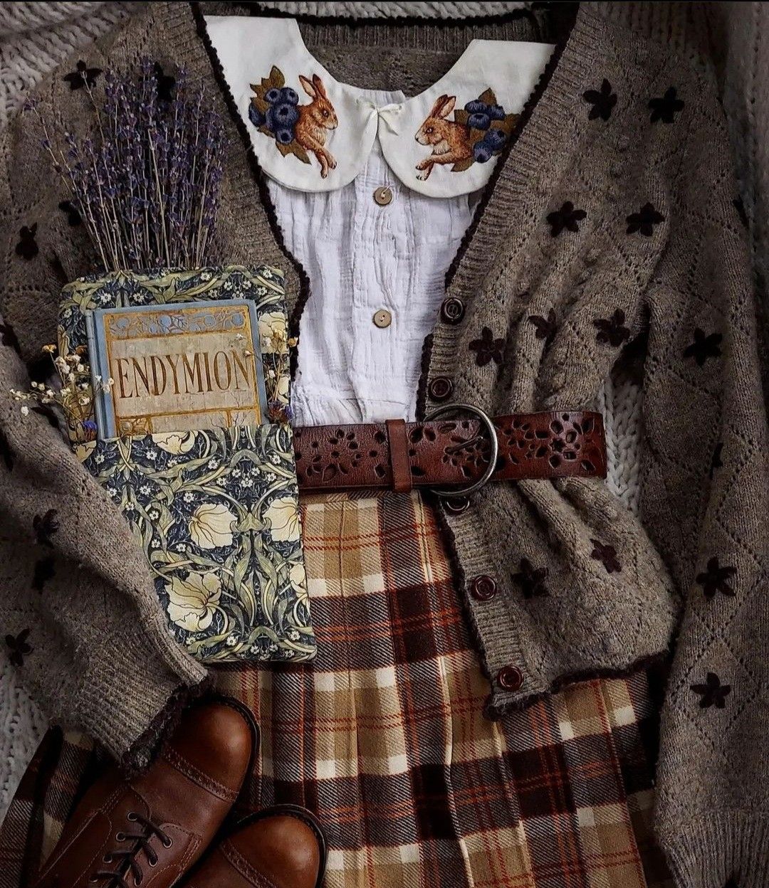 an arrangement of clothing and accessories featuring a brown cardigan with floral details, a white blouse with a rabbit and blueberrythemed collar, a brown plaid skirt, a brown leather belt, brown leather shoes, a book titled "endymion" with a floral cover, and a bundle of dried lavender and flowers.