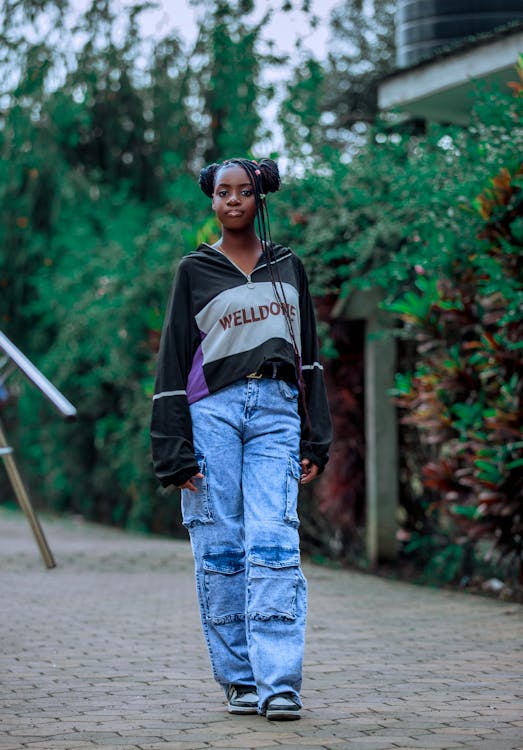 a portrait of a confident teenage african girl wearing casual streetwear. she's standing on a brick walkway, surrounded by lush greenery. she's wearing light blue denim cargo pants, a black and white oversized sweatshirt that says "welldone", and her hair is styled in braids.  the overall feeling is relaxed and stylish.