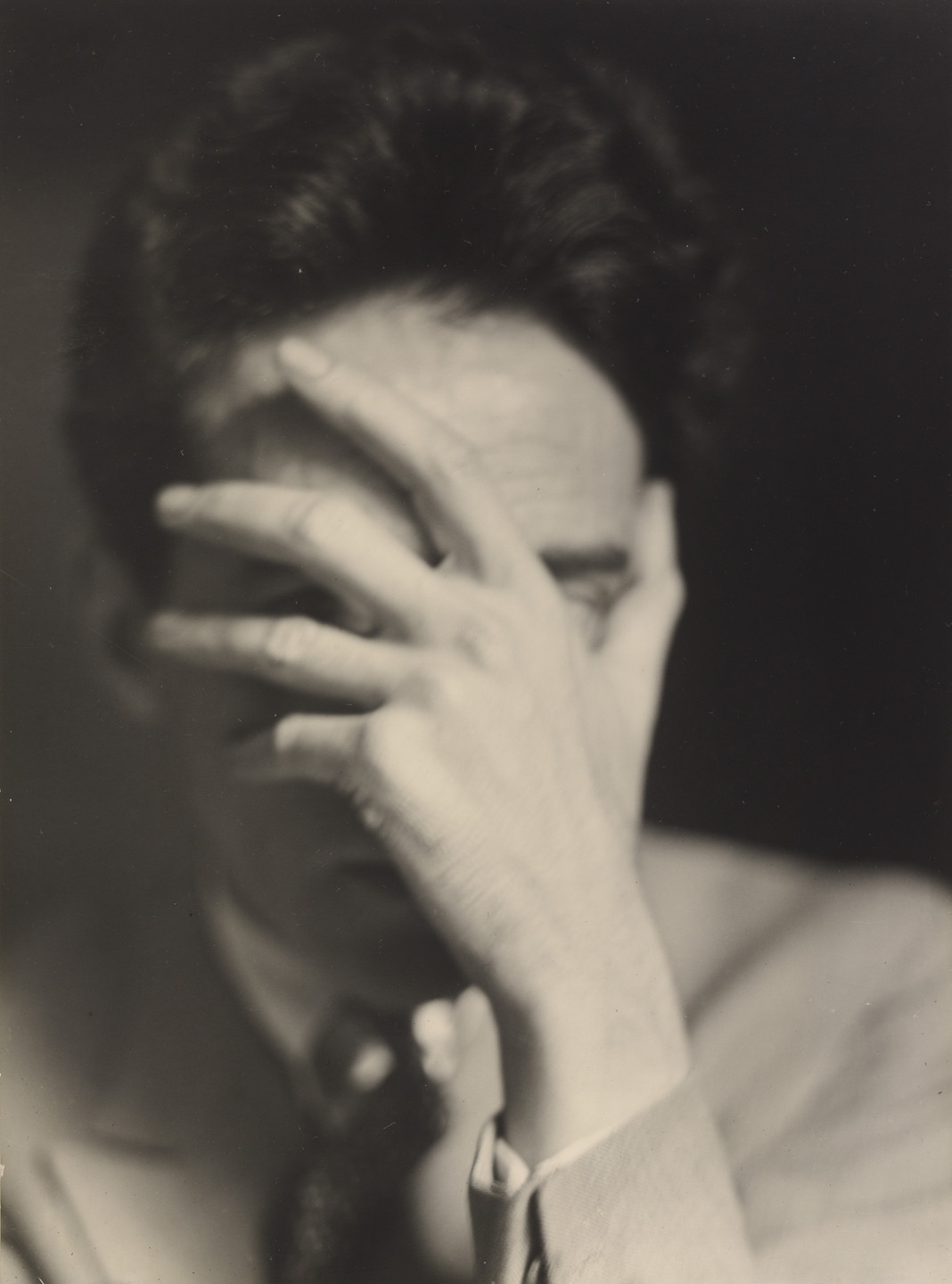 the image is a black and white closeup portrait of a man. his face is partially obscured by his hand, which is covering most of his eyes and part of his face. the lighting is dramatic, with deep shadows and highlights, creating a moody and somewhat mysterious atmosphere. the man's expression is difficult to discern, but it appears pensive or contemplative. the overall style suggests it is vintage photography from the 1930s, with artistic use of light and shadow to convey emotion.  the grain and texture of the photograph add to the vintage feel.  he is wearing a lightcolored jacket or shirt.