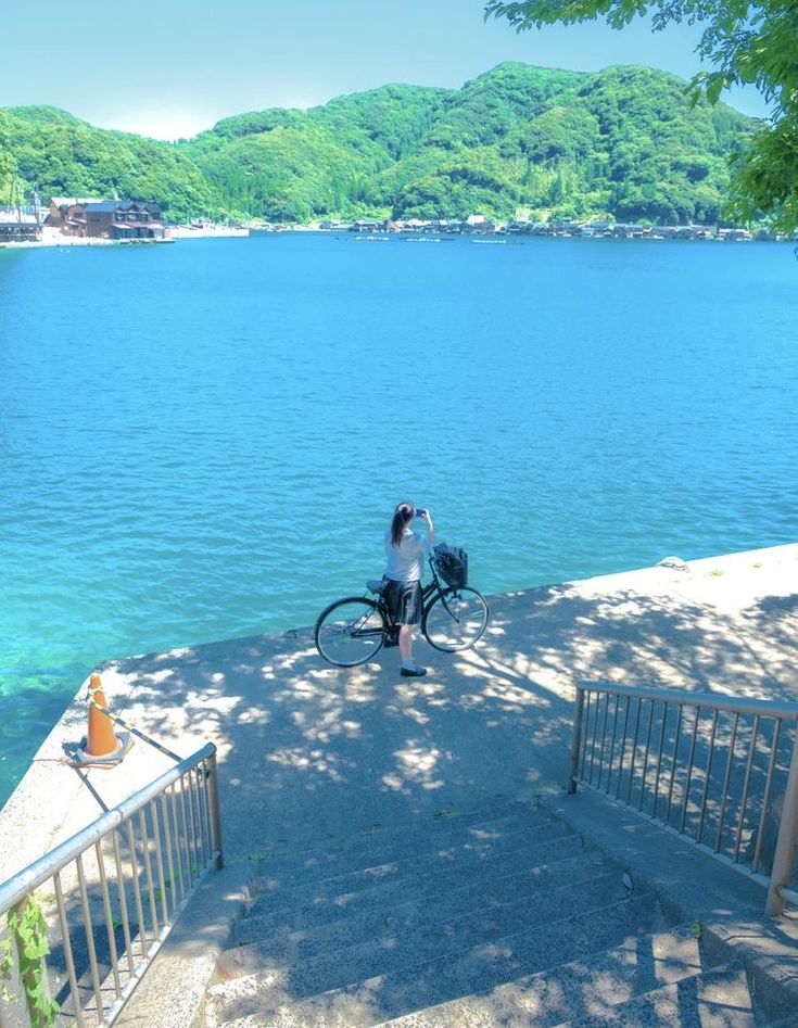 a woman with a bicycle stands on a concrete staircase overlooking a calm, blue body of water.  the water is exceptionally clear, and a picturesque scene of green hills and trees stretches across the background.  the woman appears to be taking a picture of the view.  the overall feeling is one of serenity and peacefulness.  the scene seems to be located in a coastal area, possibly japan, given the style of the houses visible in the distance.