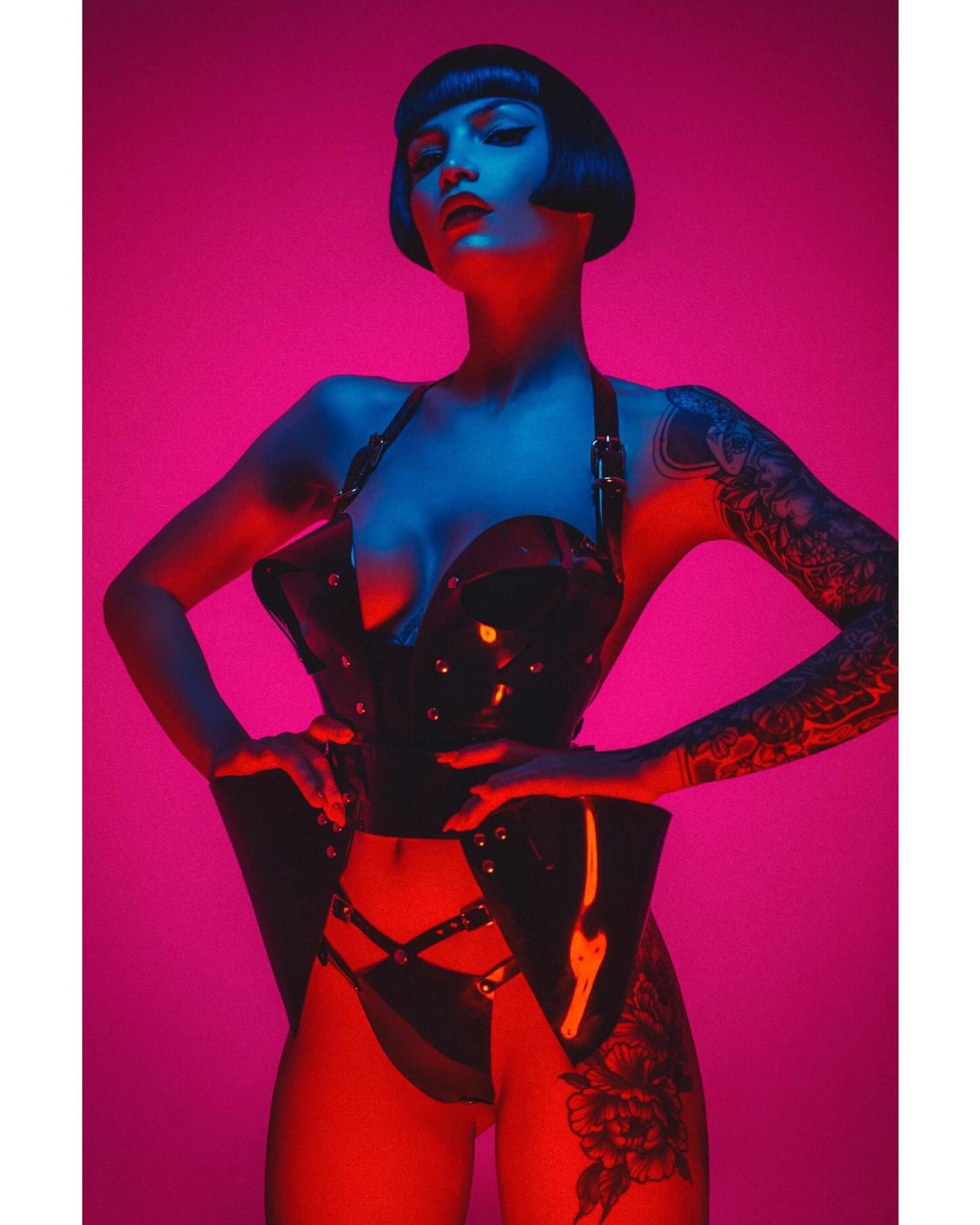 the image is a stylized studio portrait of a woman. she's wearing a black leather or pvc corset and matching lingerie that has a dark and edgy aesthetic.  the corset is structured and appears to have boning.  she has short, dark hair styled in a bob and numerous tattoos visible on her arms and legs. the background is a vibrant, contrasting pink and red neon light. the overall mood is dramatic, sensual, and evocative of alternative fashion styles like gothic or punk. the lighting creates a strong contrast and highlights the texture of the clothing and the model's features.