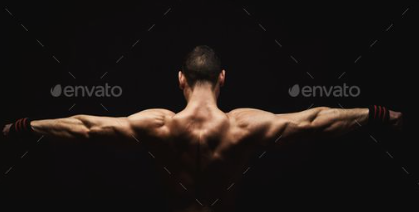 the image showcases the impressive back muscles of a muscular man. he stands with his arms outstretched, emphasizing the definition and size of his back, shoulders, and arms. the lighting accentuates his muscularity, creating a dramatic contrast against the dark background.