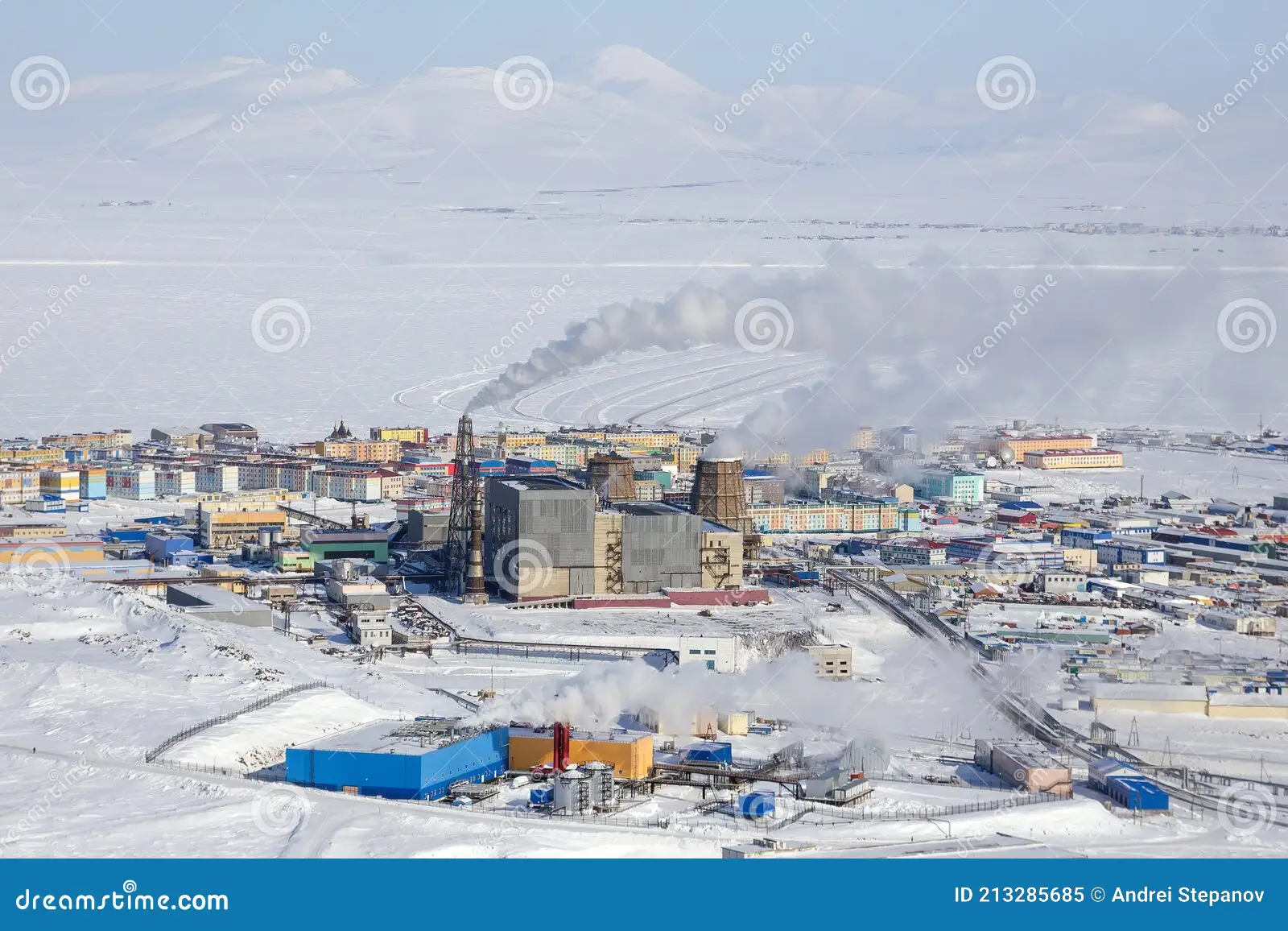 an aerial view of a city covered in snow, with a large factory belching smoke in the middle of the frame. the city is on the arctic coast of russia.
