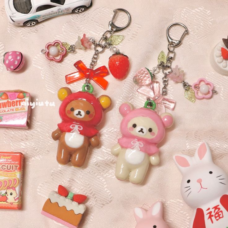 this image shows a collection of cute and kawaii items, including two keychains with bear characters (rilakkuma) wearing strawberrythemed outfits, embellished with strawberry and floral charms, alongside other toys and trinkets such as a car toy, bunny figurines, a candy box, and dessert figurines on a textured pink background.