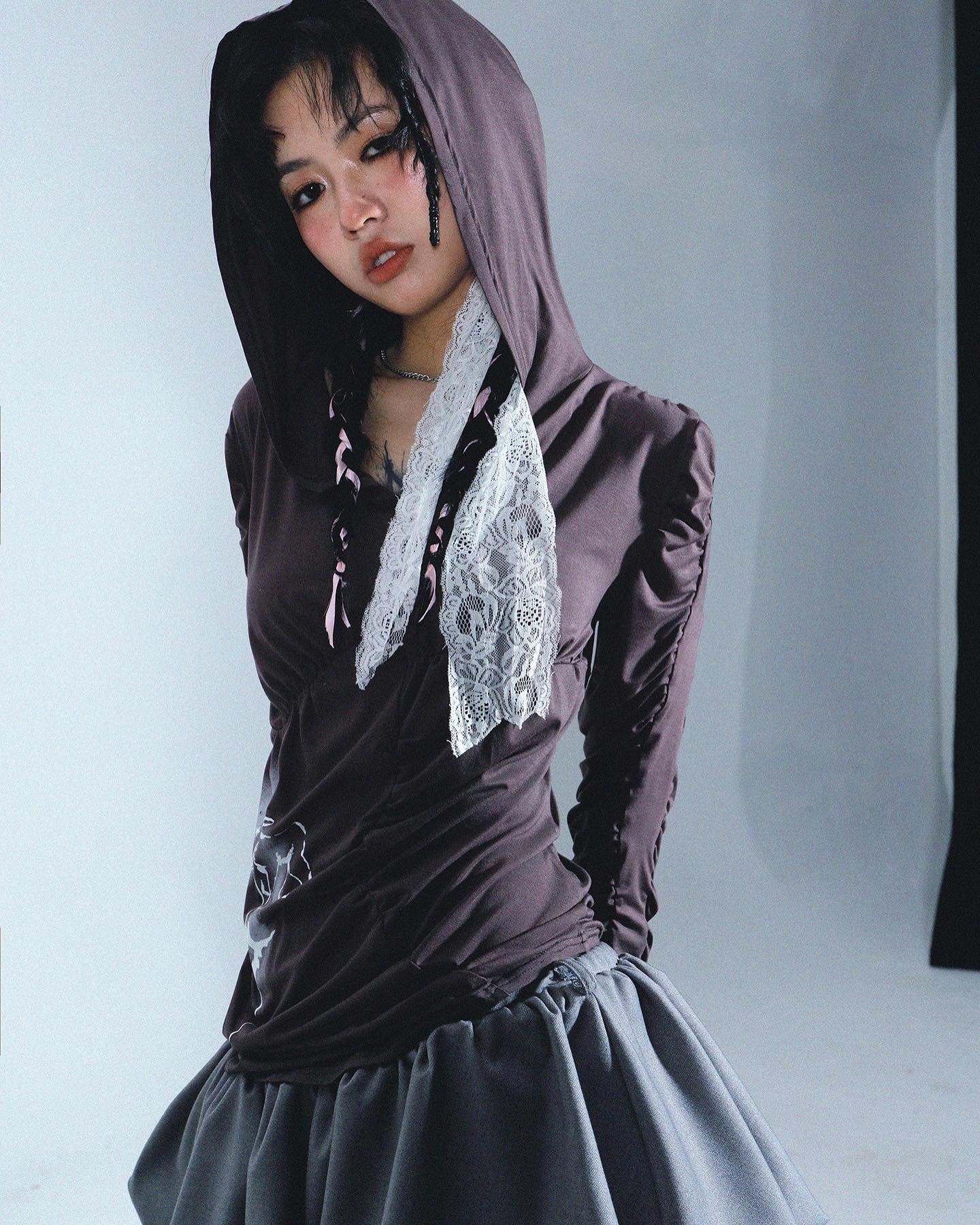 a closeup shot of a young woman modeling a dark brown hooded longsleeved top with lace and ribbon details, paired with a grey pleated skirt. she is posing against a light grey backdrop. the overall style is edgy and alternative, incorporating elements of dark academia and gothic aesthetics. her makeup is subtle but noticeable, complementing the dark clothing. the image is welllit and professionally taken, likely part of a fashion photoshoot.