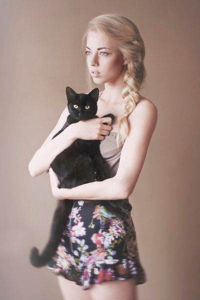 a young woman with blonde hair in a braid holds a black cat in her arms. she is wearing a floral print skirt and a nudecolored top. the background is a muted beige.