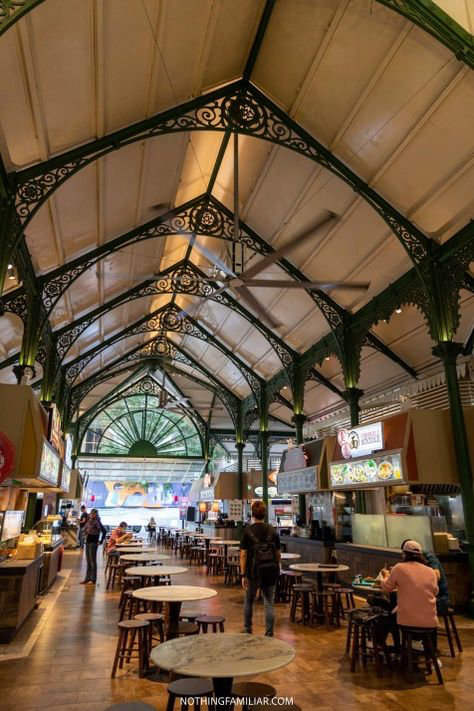 the image shows the interior of lau pa sat (telok ayer market) in singapore. the focus is on the beautiful victorianera cast iron architecture of the building's ceiling and the food stalls and tables below.  people are seated at tables, eating and enjoying the atmosphere of the bustling hawker center. the image captures a blend of historical architecture and modernday food culture. the lighting is relatively subdued, enhancing the architectural details.