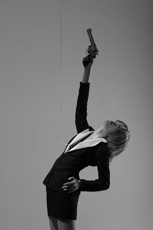 a black and white image of a woman in a black business suit. she is holding a revolver in her raised right hand, her body arched in a dynamic pose. a thin line extends from the gun to the upper left, suggesting a connection or manipulation. the background is a simple gray, focusing attention on the subject. the overall mood is dramatic and suggestive of power, danger, and control. the image is stylish and artistic, with strong contrasts and clean lines.
