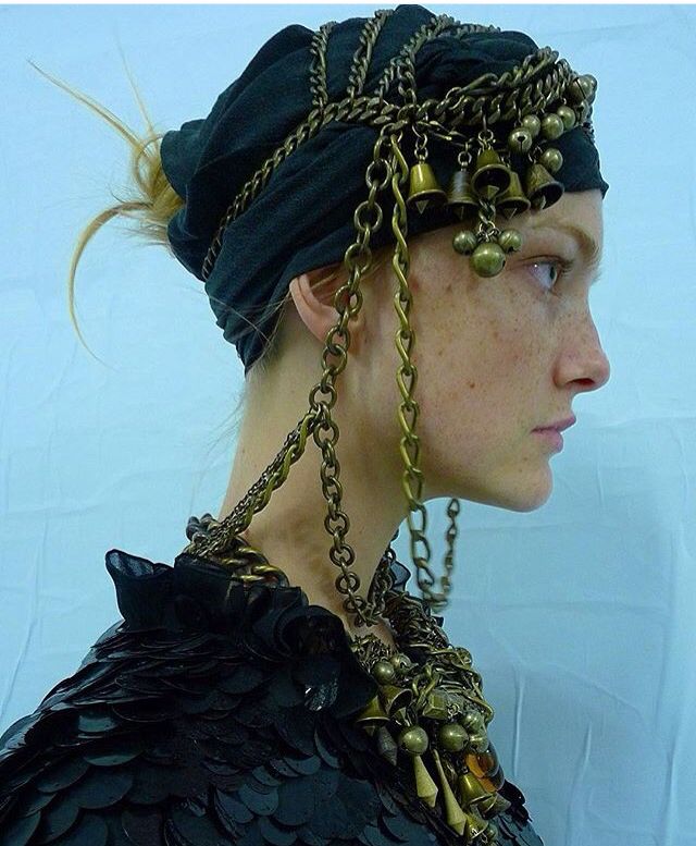 a woman with freckles is shown in profile, wearing a black turban adorned with heavy, layered gold chains and bells. she is also wearing a black shoulder covering made of overlapping round pieces, similar to scales. the background is a light blue.