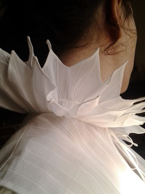 a closeup shot shows the back of a person's head and neck, adorned with a large, white ruff or collar made of a sheer, textured fabric. the collar has a petallike design, extending outwards from the neck. the person's ear is visible, and dark hair is loosely pulled back. the garment beneath the collar is also made of a similar sheer, white fabric with subtle lines.