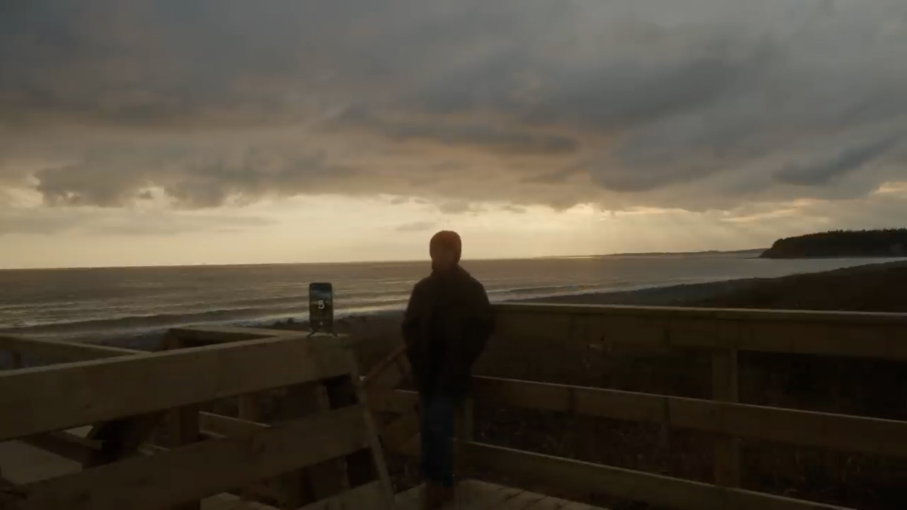 a man stands on a wooden boardwalk overlooking a beach and the ocean. the sun is setting in the distance, casting a warm glow on the clouds and water. the sky is a mix of dark and light clouds, creating a dramatic backdrop. the man is silhouetted against the light, and he appears to be looking out at the view. a phone is mounted on a tripod in front of him, suggesting he may be taking a picture or video.