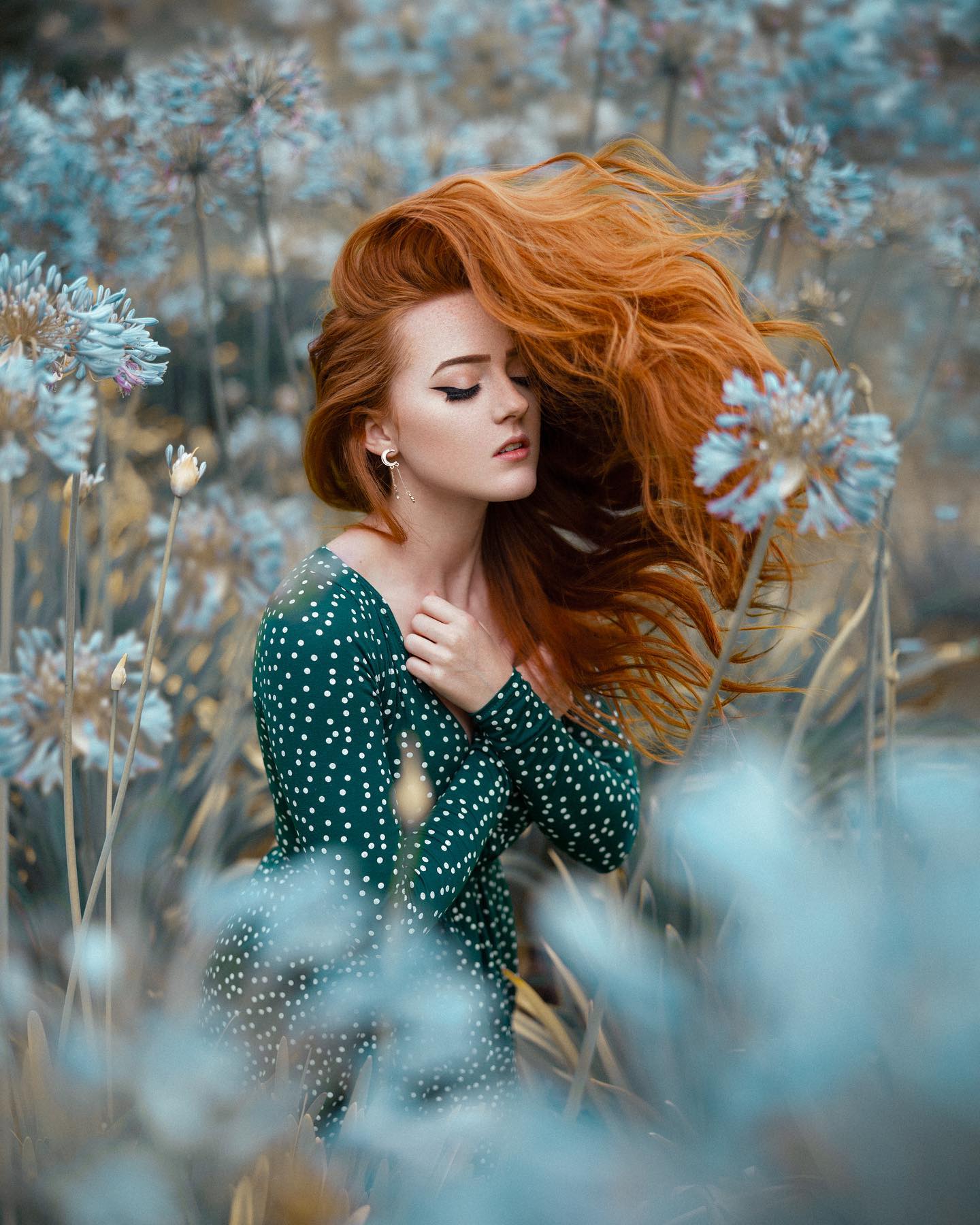 a beautiful redhead woman in a green dress, stands amongst blue and white wild flowers. the photo is taken at eye level, and the soft lighting creates a dreamy atmosphere. the wind is blowing through her hair, which adds to the overall ethereal feel of the image.
