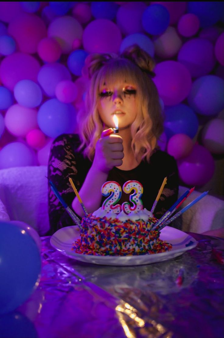 a woman is sitting in front of a birthday cake with candles that spell out "23". she is holding a lighter and about to light the candles.  she has a happy look on her face and is surrounded by colorful balloons. the background is a wall of purple balloons and the table is covered in a sparkly tablecloth.
