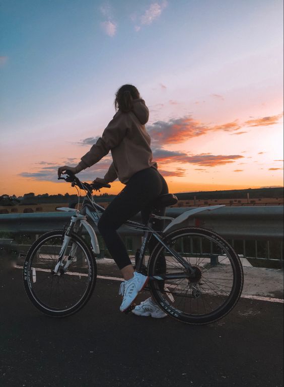 a girl in black leggings and a brown hoodie is sitting on her silver and black mountain bike on the side of a road. the road goes from the left to the right side of the image. behind her is a field, and in the background, there is a beautiful sunset with orange, yellow, blue, and purple clouds.  she is looking towards the right side of the image towards the sunset.