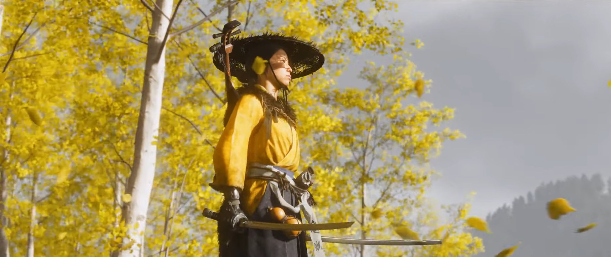 a woman in a yellow robe, wearing a hat and carrying a katana, stands in a forest of yellow trees. the trees are bare and the leaves are falling. the sky is grey. she is looking straight ahead. she appears to be a character from a video game.