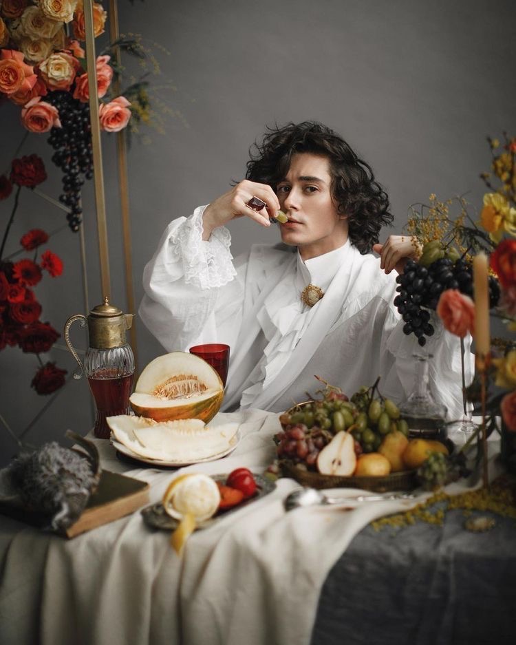 a young man with curly hair, dressed in a white, ruffled shirt reminiscent of renaissance fashion, sits at a table laden with food and flowers. he holds a grape to his lips, his gaze fixed on the viewer. the scene is reminiscent of a classical still life painting, with a muted color palette and soft lighting, further emphasized by the arrangement of fruit, flowers, and a vintage decanter.