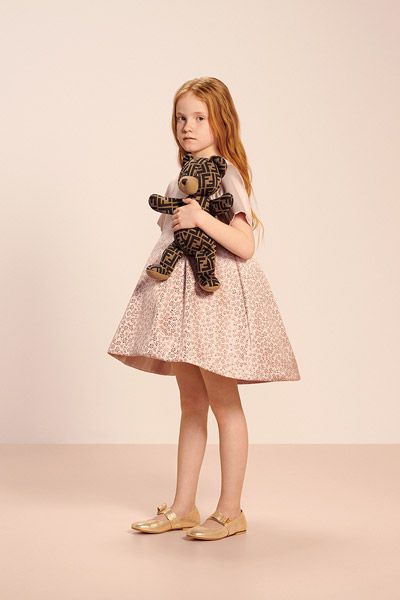 a full shot of a young girl in a pink floral dress, holding a fendi teddy bear. she is wearing gold shoes and is posing against a light beige background. the overall aesthetic is sophisticated and charming.