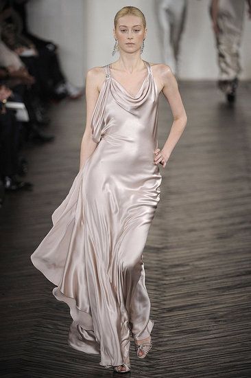 a runway model walks the catwalk wearing a pale pink satin gown with a cowl neck and strappy sandals. she has blonde hair pulled back and long, dangly earrings. the background shows blurred figures and a wooden floor.