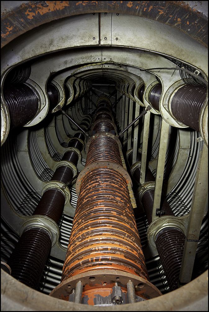 the image is a detailed closeup view of the interior of a large, cylindrical electrical transformer.  the central focus is on a massive, rustcolored coil made up of numerous concentric windings.  surrounding this central coil are other coils of a darker, almost black color, all housed within a circular metallic structure showing signs of age and wear, including rust and paint discoloration. the overall impression is one of immense scale and complex engineering. the image is taken from a perspective looking directly down into the center of the transformer.