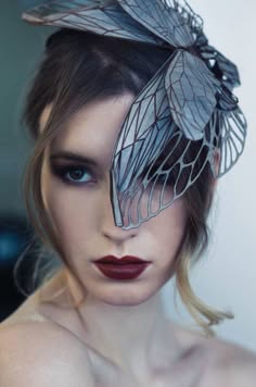 a woman with long, dark hair is wearing a cicada wing as a headpiece. she is looking at the camera and has on dark eye makeup and a dark red lipstick. the cicada wing is covering half of her face and has a delicate and intricate design. the woman is standing in front of a white background.
