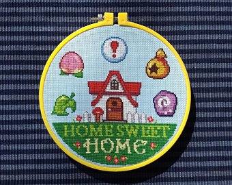 a closeup shot of an animal crossing crossstitch project, featuring a house, a leaf, a bell bag, and a home sweet home inscription. the crossstitch is surrounded by a yellow hoop and is set against a blue and white striped fabric.