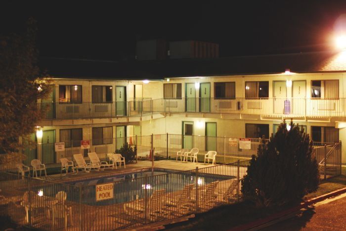 a night view of a motel with a swimming pool in the front. the pool is surrounded by a fence and there are chairs set up around it. there are lights on the outside of the motel building.