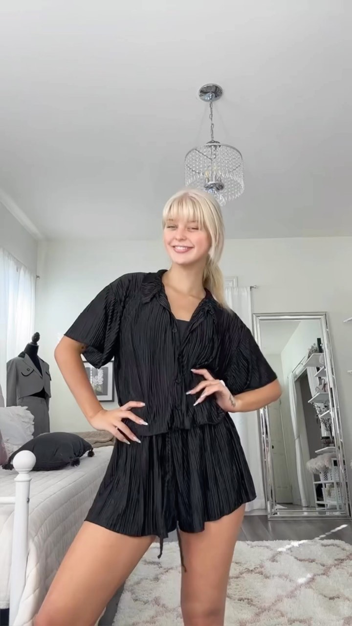 a woman stands indoors in a bedroom, wearing a black blouse and shorts set. a mirror stands in the background.