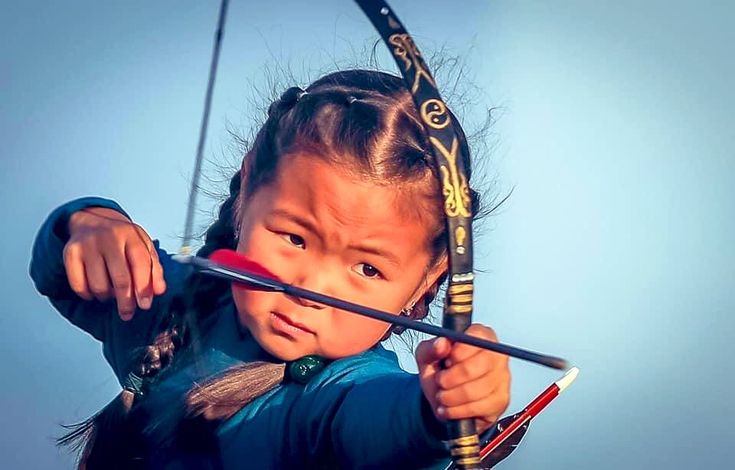 a young girl dressed in blue clothing is drawing a bow and arrow, focusing intensely on her target. she has braids in her hair and a serious expression on her face. the bow is ornately decorated. the image evokes a sense of tradition and skill in archery.