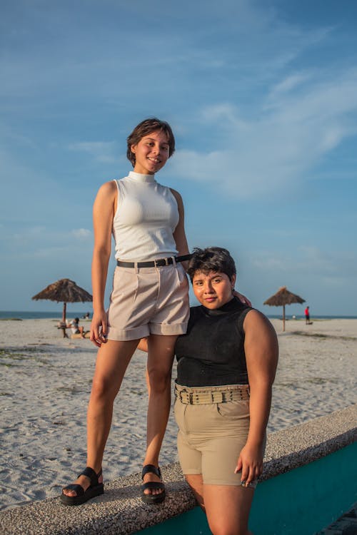 two young women are standing on a sandy beach, smiling at the camera. they are both wearing casual summer clothing. the taller girl is wearing a white sleeveless top and beige shorts and the shorter girl is wearing a black top and khaki shorts.  the beach has a few small palmthatched umbrellas in the background and the sky is a clear blue. they appear to be enjoying a sunny day at the beach.  the overall feeling of the image is happy, carefree, and joyful.