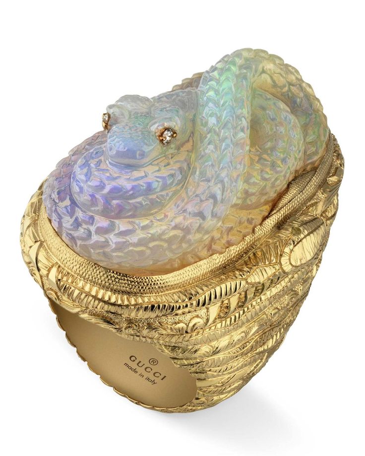 a closeup shot of a gucci ring. the ring features a golden band with intricate patterns, topped with a coiled snake made of opal material. the snake has small gemstone eyes. the inside of the ring is labeled "gucci made in italy".