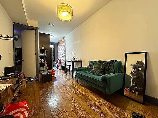 a view of a living room in a small apartment. the room has a green couch, a wood floor, and a brick wall. there is a table with a chair near the couch, a bookshelf in the corner, a tv on a stand, and a mirror on the wall. the room is decorated with a lamp, a rug, and some pillows.