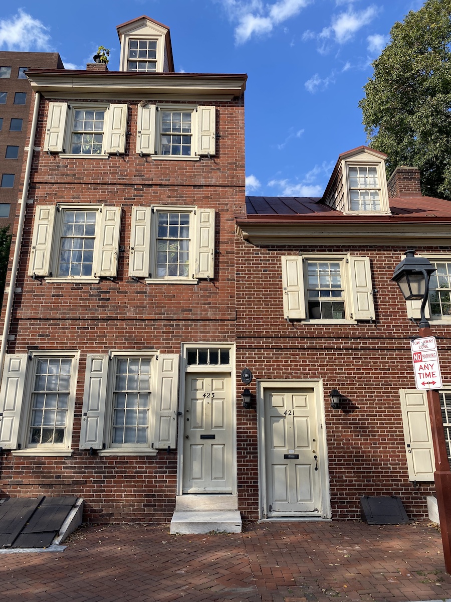 the image shows a red brick building with a series of windows and shutters. the building has two doors, each with a house number (423 and 421). there is a streetlamp and a "no parking any time" sign in front of the building. the building's architecture includes dormer windows on the roof.