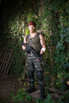 a man dressed in camouflage fatigues and tactical gear stands in a lush jungle setting, holding a rifle. he is wearing a red bandana and knee pads.  the scene is dark and moody, suggesting a military or action setting. the man appears to be posed for a photograph.