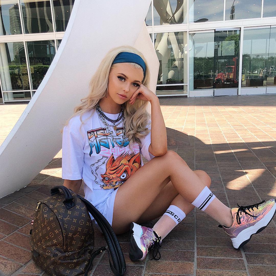 a young woman with blonde hair sits on a brick pavement with a backpack next to her. she is wearing a white tshirt with a graphic print, shorts, socks with text, and sneakers. she also has a blue headband and several necklaces. she is leaning her head on her hand. the background includes modern architecture and a sunny sky.