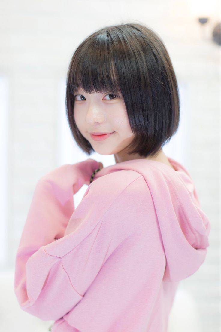 a young asian woman with short, black hair and bangs is wearing a pink hoodie and is smiling slightly as she looks over her shoulder. the background is white. she is indoors.