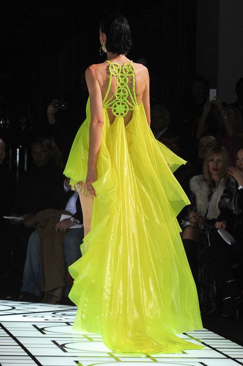 a fashion model walks the runway in a striking neon yellow dress with a sheer, flowing train. the dress features a delicate, intricate lace pattern on the back, creating a captivating silhouette. the model's dark hair and makeup add to the drama of the look. the runway is lit up by a bright spotlight, highlighting the dress's vibrant color and the model's confident pose. the backdrop is a blurred sea of spectators, emphasizing the allure of the fashion world.