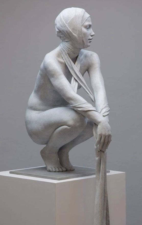 a white plaster sculpture of a woman, sitting with her legs bent at a 90degree angle, with a piece of draped cloth covering her head and body. the sculpture is sitting on a white pedestal. it appears to be a contemporary work of art.