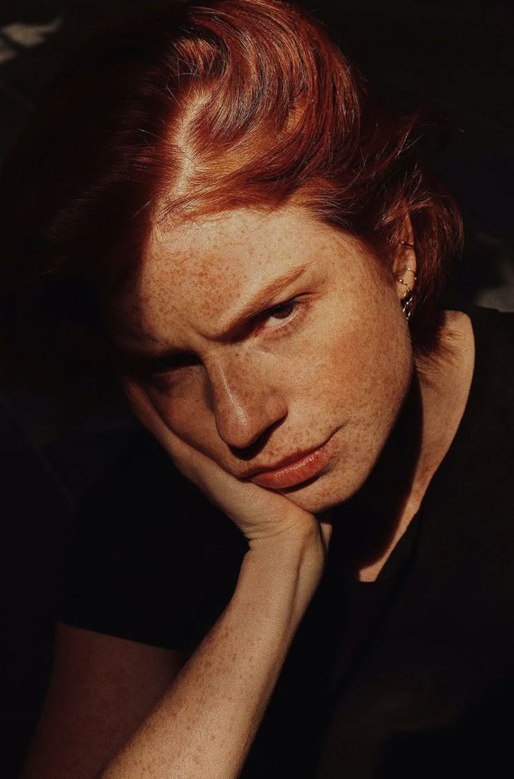 a closeup portrait of a woman with red hair and freckles resting her chin on her hand. she has a serious, pensive expression. the lighting is dramatic with strong shadows across her face. she is wearing a black shirt, and her ear has multiple piercings.