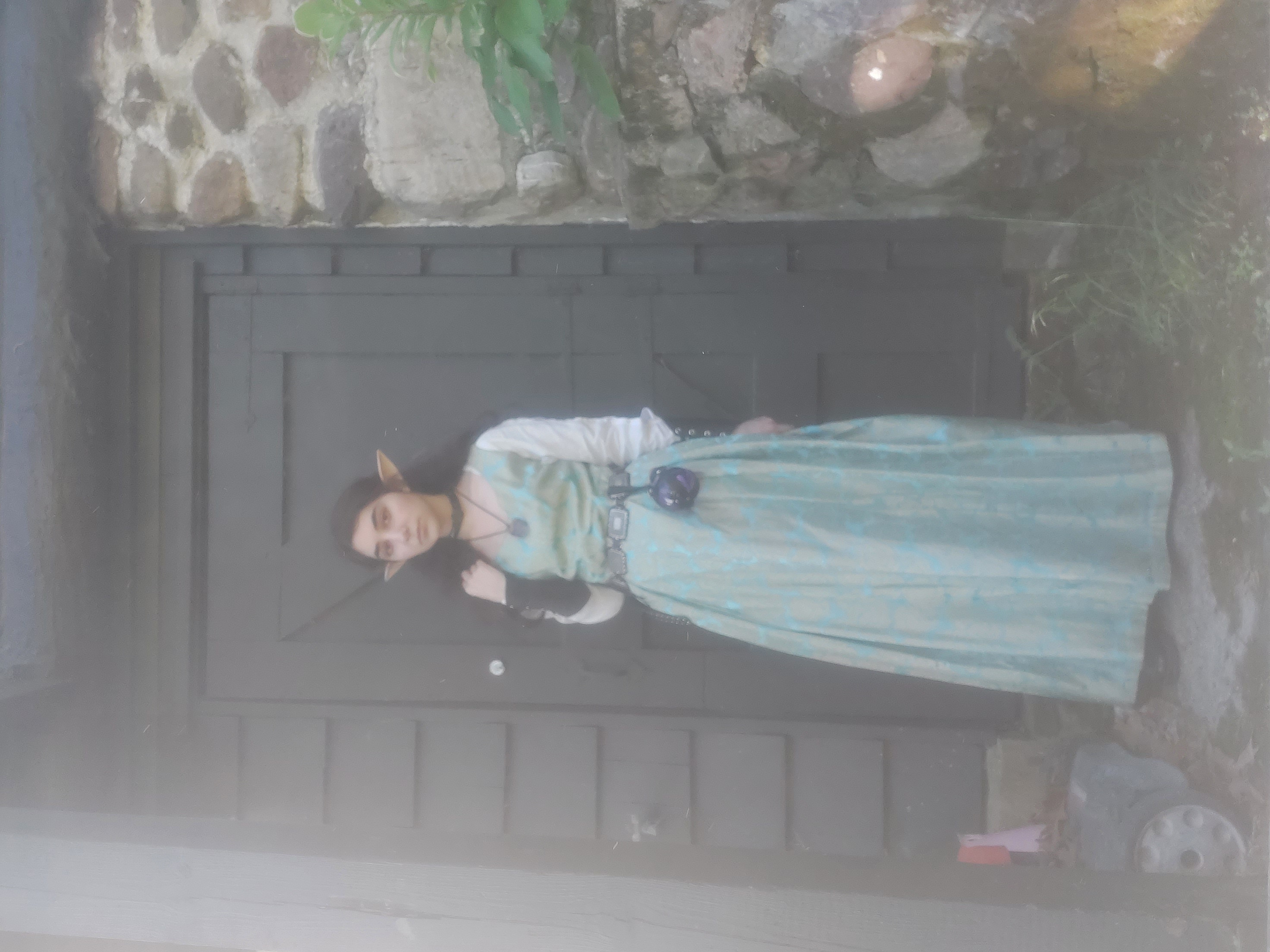 a young woman dressed in an elf costume stands in front of a stone wall and dark wooden door. her costume includes a long, flowing teal gown with a subtle pattern, a dark belt, and a necklace with a pendant. she is wearing prosthetic elf ears. the overall aesthetic is whimsical and mystical, suggestive of a fantasy setting.  the photo has a slightly soft and dreamy focus.