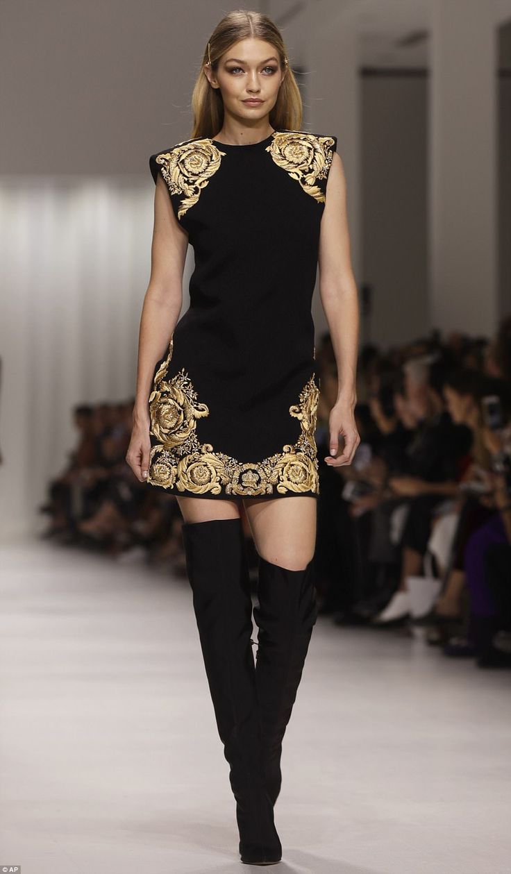 gigi hadid walks the runway at a versace fashion show. she is wearing a short black dress with gold embroidery and tall black boots. the background shows the audience watching the show. the image is welllit and professionally taken.