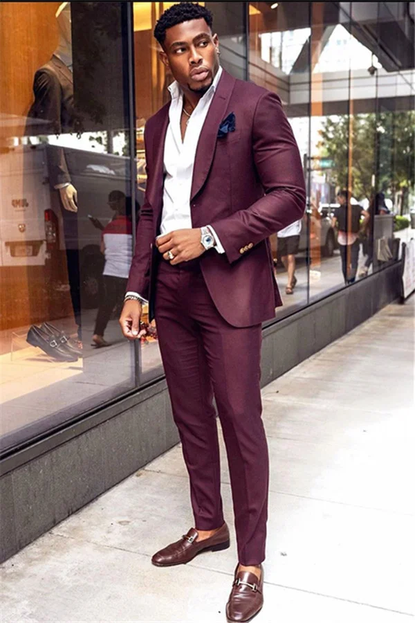 a welldressed african american man stands on a city street wearing a sharp, slimfitting burgundy suit. he pairs the suit with a crisp white shirt, brown loafers, and a watch. the overall look is stylish, elegant, and sophisticated, showcasing a modern take on men's formal wear.  the background includes a storefront window reflecting his image and other passersby.
