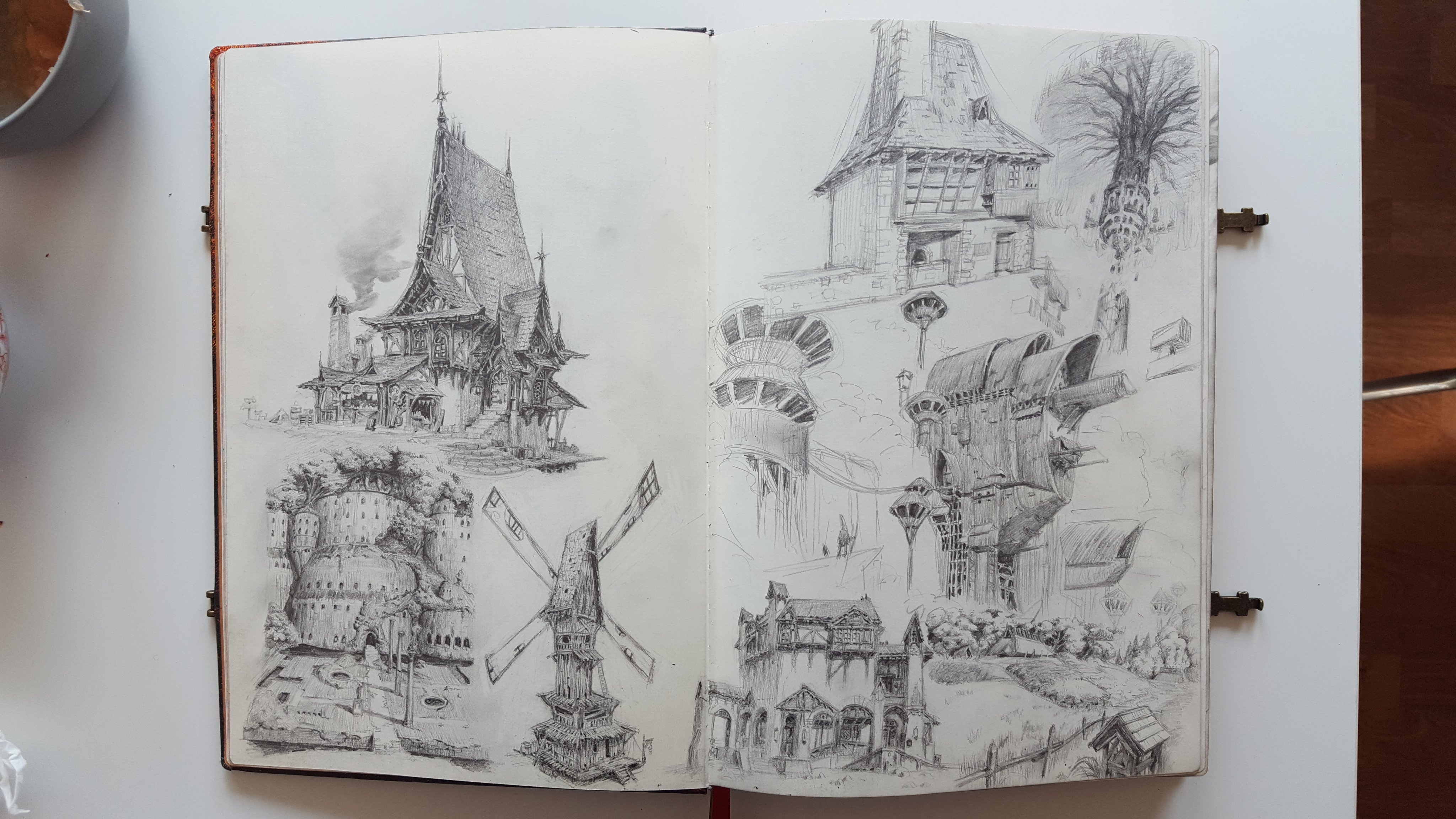 an open sketchbook lies on a table, showcasing detailed pencil sketches of fantastical buildings and windmills. the drawings exhibit a range of architectural styles and perspectives.