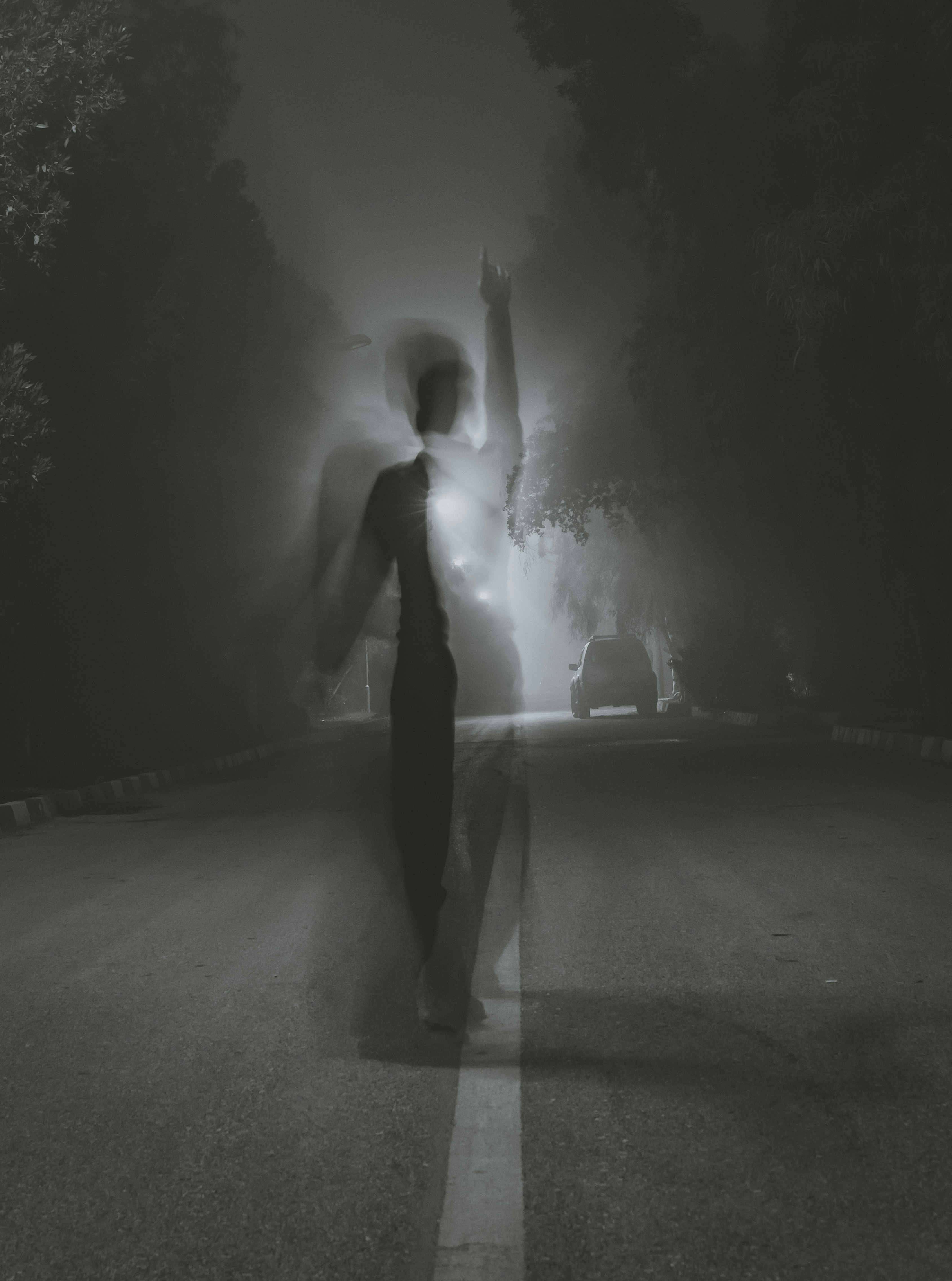 a black and white photo of a man standing in the middle of an empty street at night, with his arm raised. the photo is taken from a low angle, and the man is silhouetted against the headlights of a car in the distance. the photo is blurry and has a dreamlike quality.