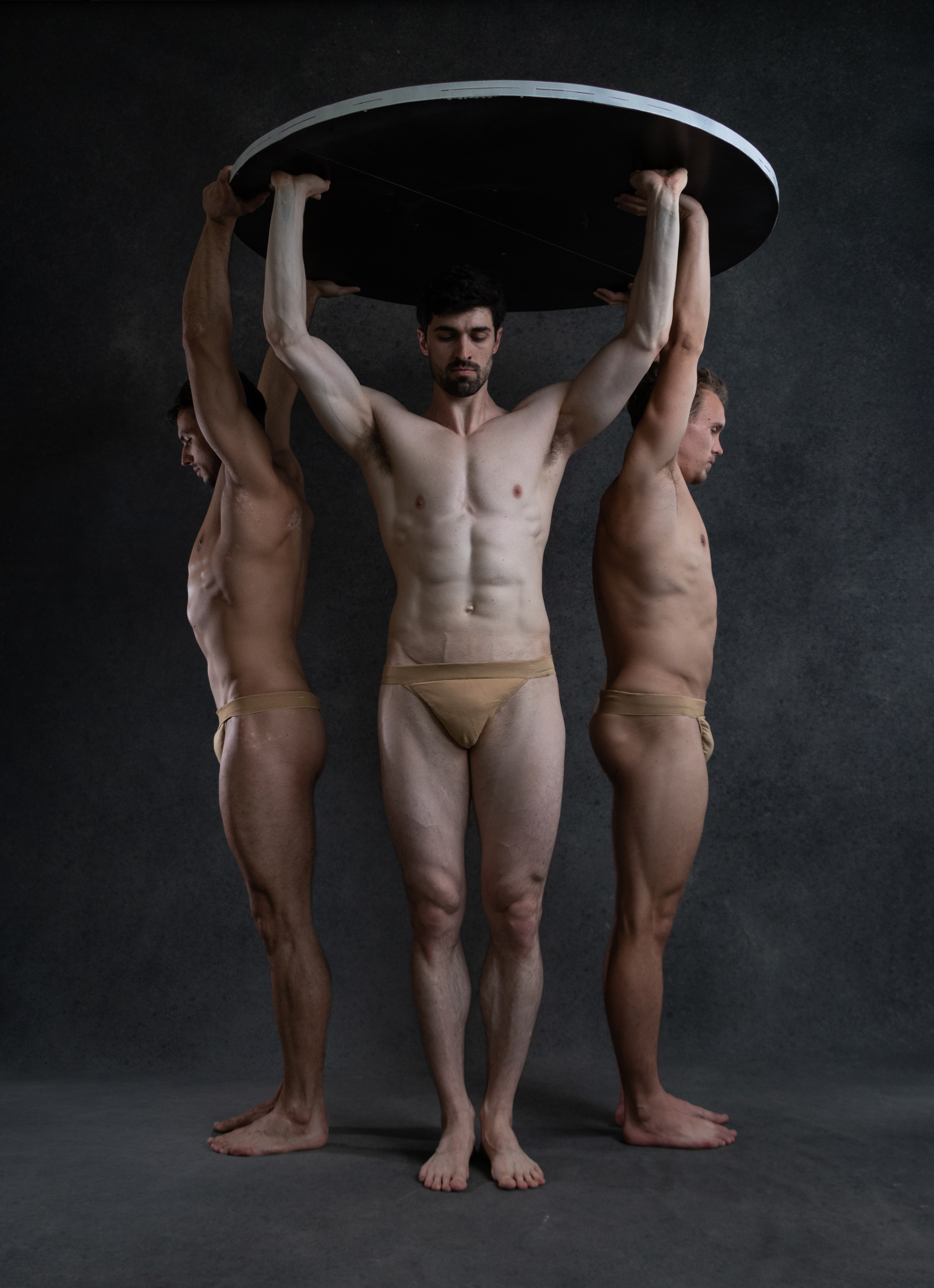 he image showcases three muscular men in a studio setting. they are positioned against a dark gray background, illuminated by soft, even studio lighting. two of the men stand on either side, their bodies facing outwards and slightly angled towards the viewer. they each hold an edge of a large, black circle above their heads. the third man stands in the center, facing forward, with his arms raised, supporting the circle from beneath. all three men are nude and adopt a stoic expression, their muscular physiques accentuated by the lighting and pose. their bodies create a sense of strength, balance, and symmetry within the composition.