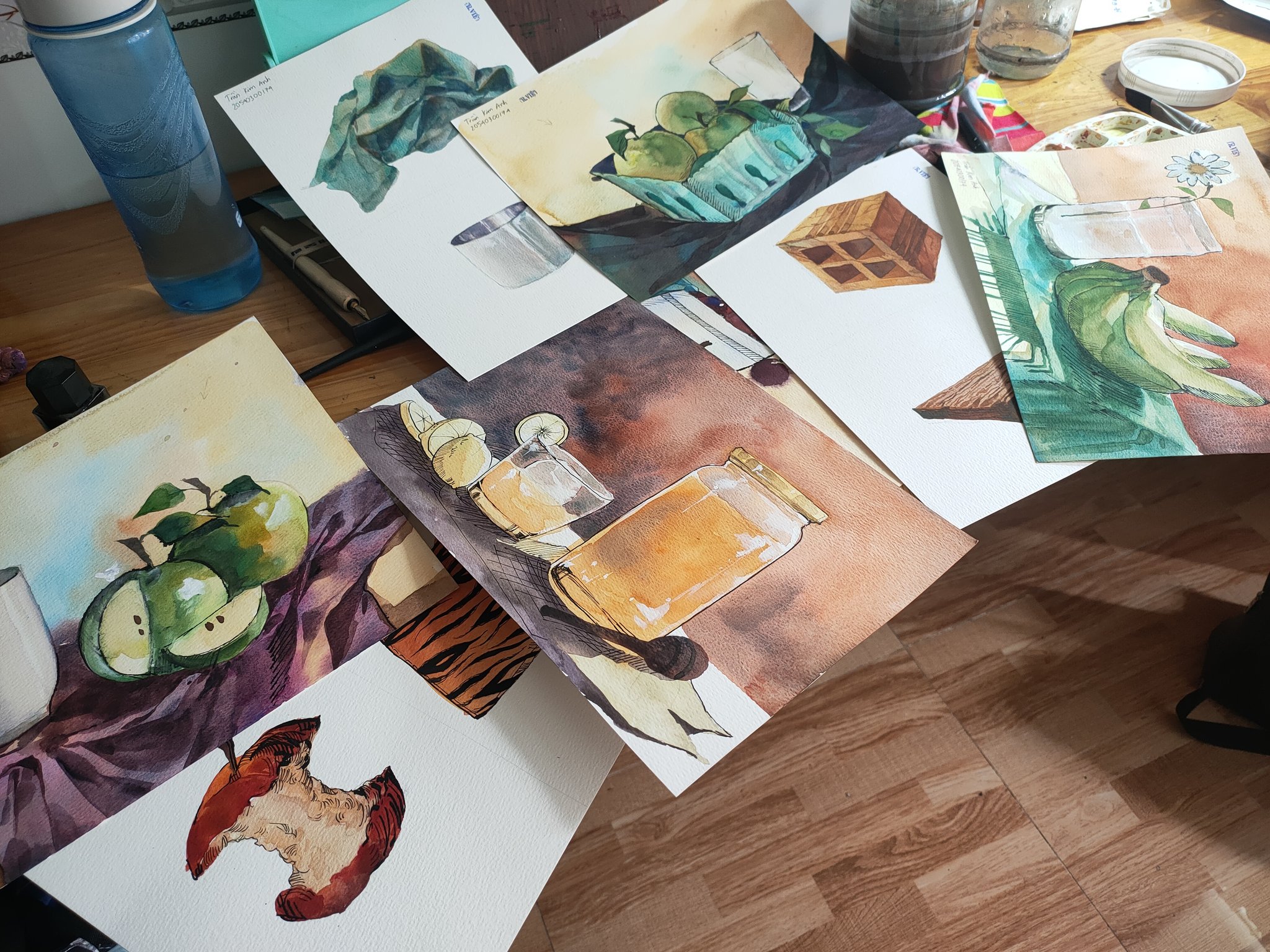 the image shows a collection of watercolor paintings arranged on a wooden table.  the paintings depict various still life subjects, including apples, bananas, a jar of what appears to be honey or juice, glasses, and other objects.  some paintings are more complete than others, suggesting a work in progress. the overall setting appears to be an art studio or workspace, with art supplies visible in the background.  the style is realistic, with attention to detail in the rendering of textures and light.  the paintings are on individual sheets of paper, and some show notations or labels.