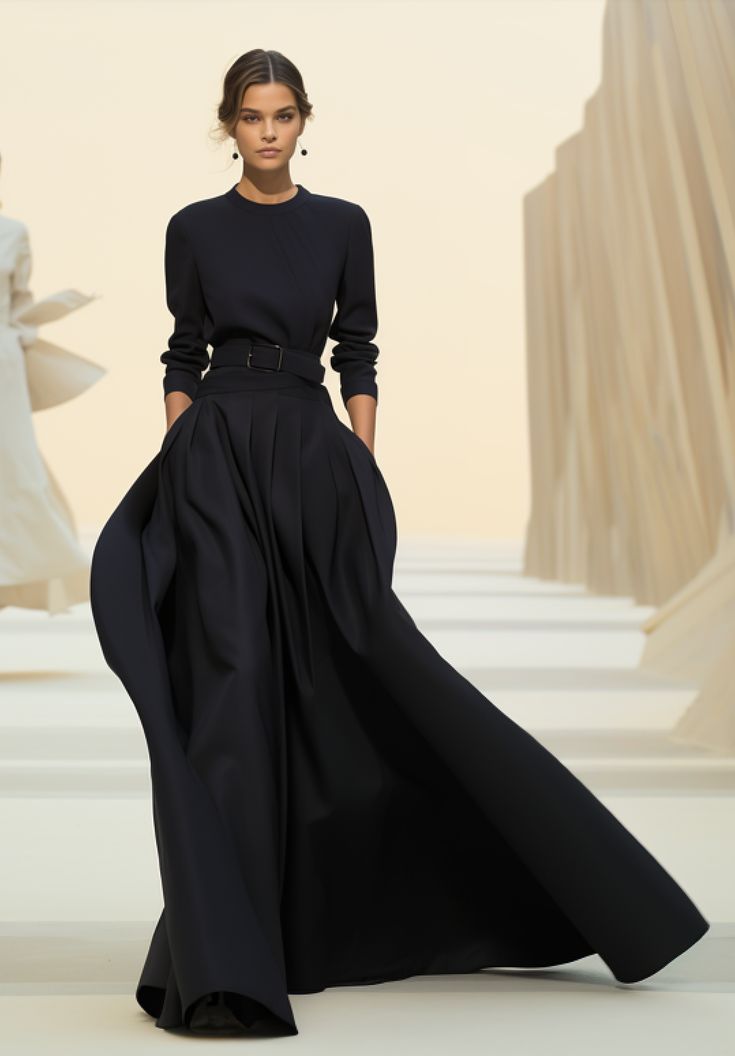 a woman stands on a runway wearing a longsleeved black dress with a belted waist and a dramatically pleated skirt. her hands are casually in her pockets. the runway has a minimalistic backdrop.