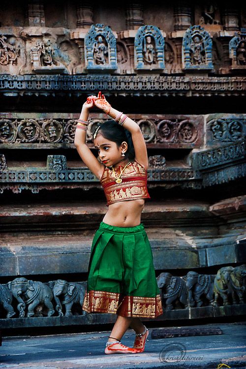 a young indian girl dressed in traditional bharatanatyam attire is captured in a dance pose in front of an intricately carved temple. she wears a vibrant red and gold cropped top, a green skirt with a gold border, and adorned with jewelry including bracelets, earrings, a necklace, and anklets. her hands feature henna patterns, and she holds her arms raised in a classic dance gesture. the backdrop is an ancient temple with detailed architectural carvings and sculptures, adding to the cultural and artistic context of the image.